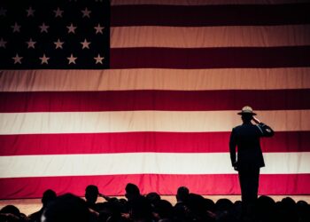 man standing on stage facing an american flag
