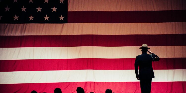 man standing on stage facing an american flag